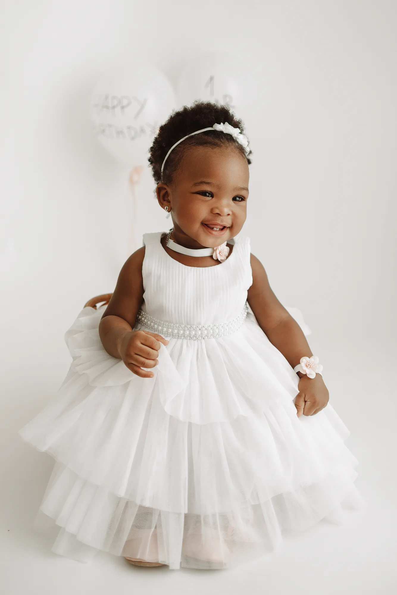One-year-old girl smiling in a white tulle dress in front of birthday balloons