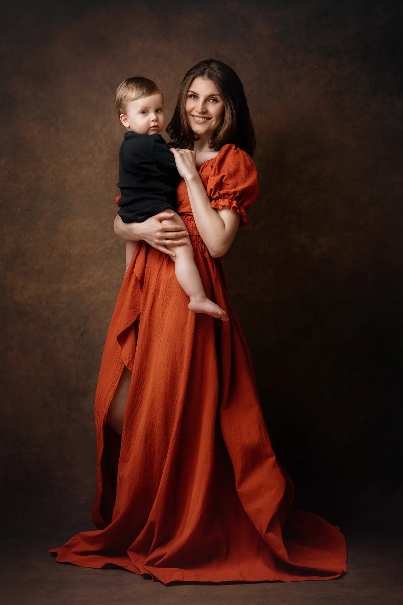 Mother in a rust-red gown holding her baby son, fine art family portrait