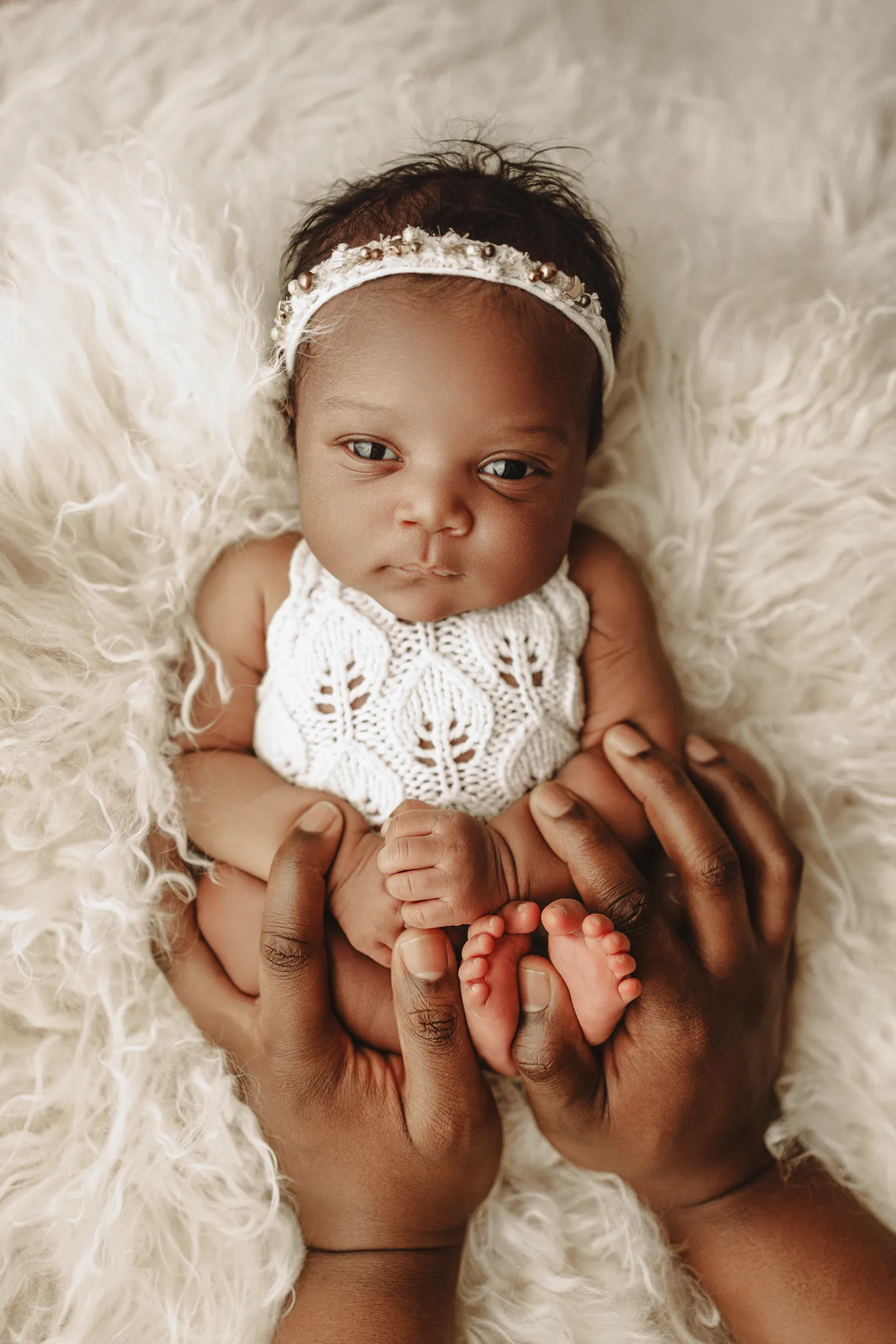 Newborn baby girl in a white knit romper and headband, held by parent's hands on cream fur