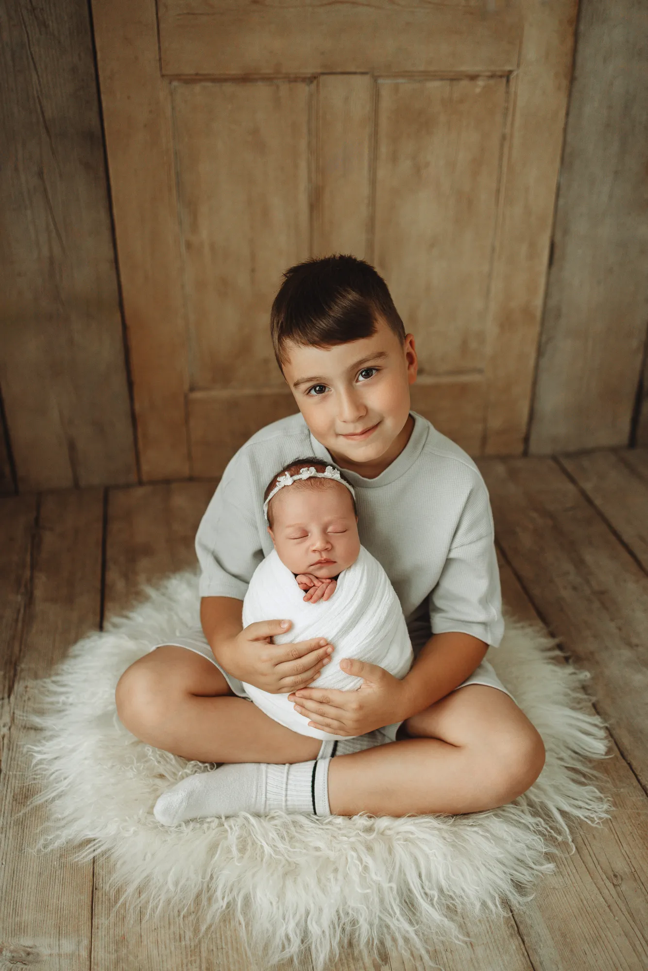 Big brother sitting on a sheepskin rug cuddling his swaddled newborn sister