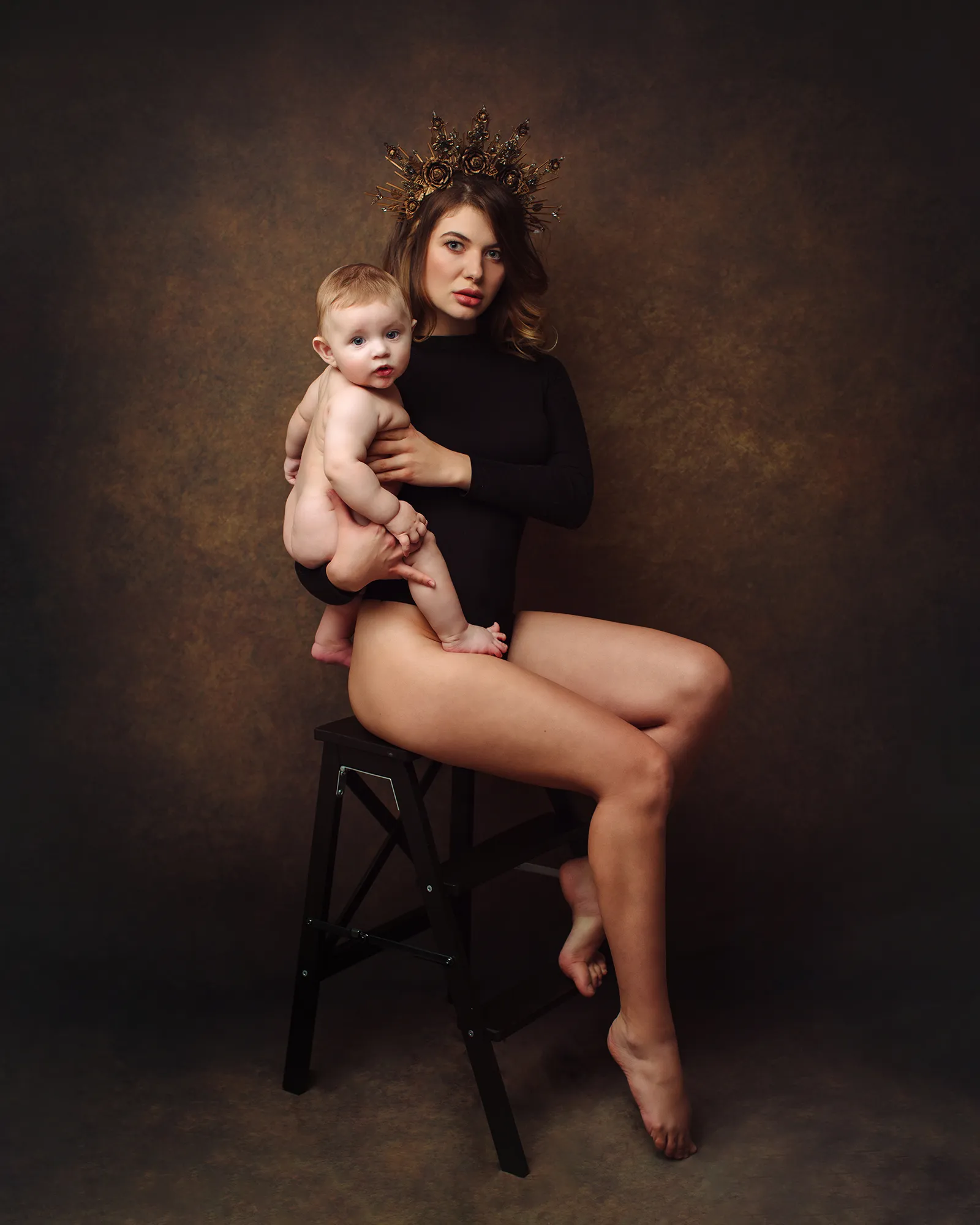 Mother in a black bodysuit and gold crown holding her baby on a stool