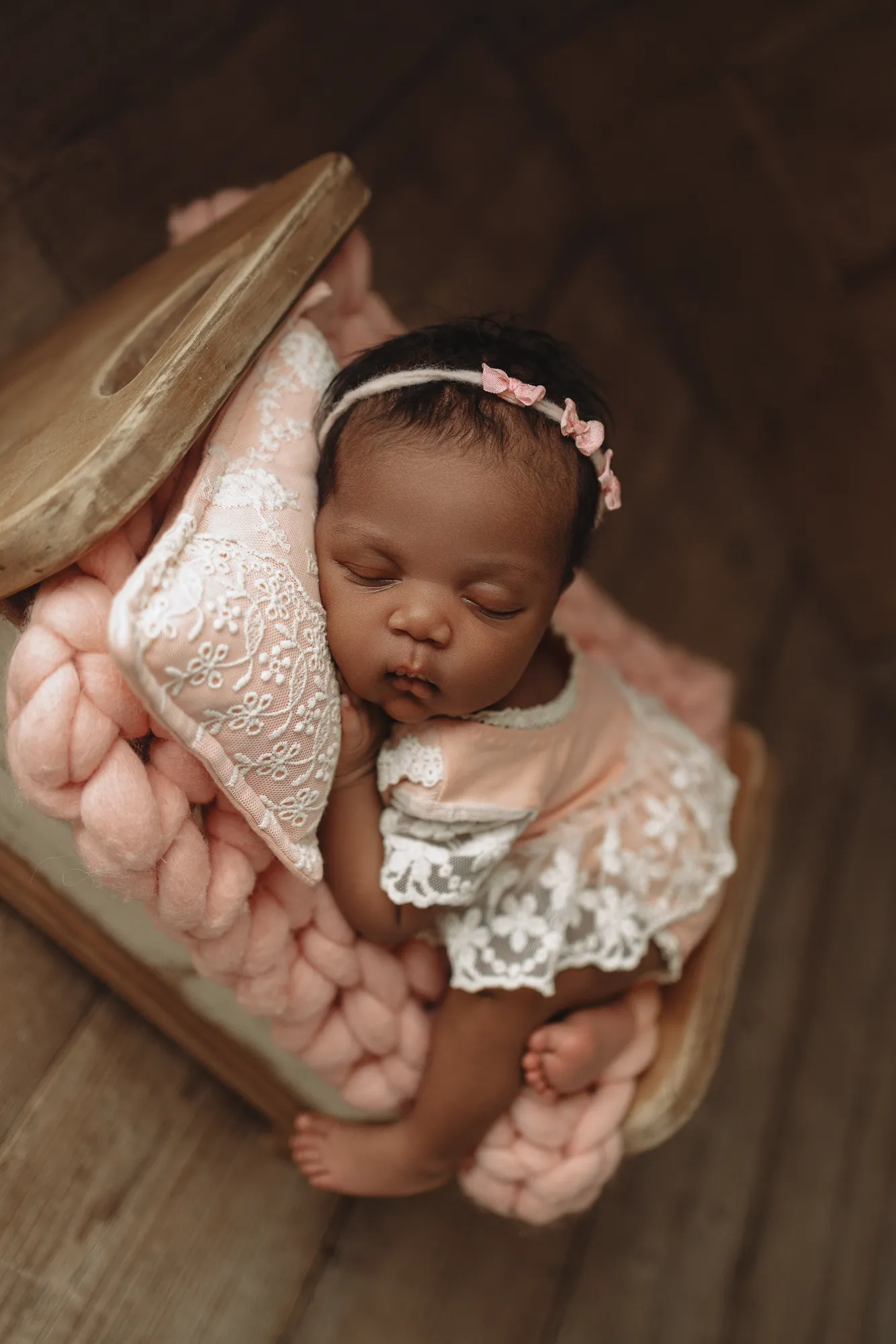 Newborn asleep on a pink knit blanket in a wooden bed with a lace pillow