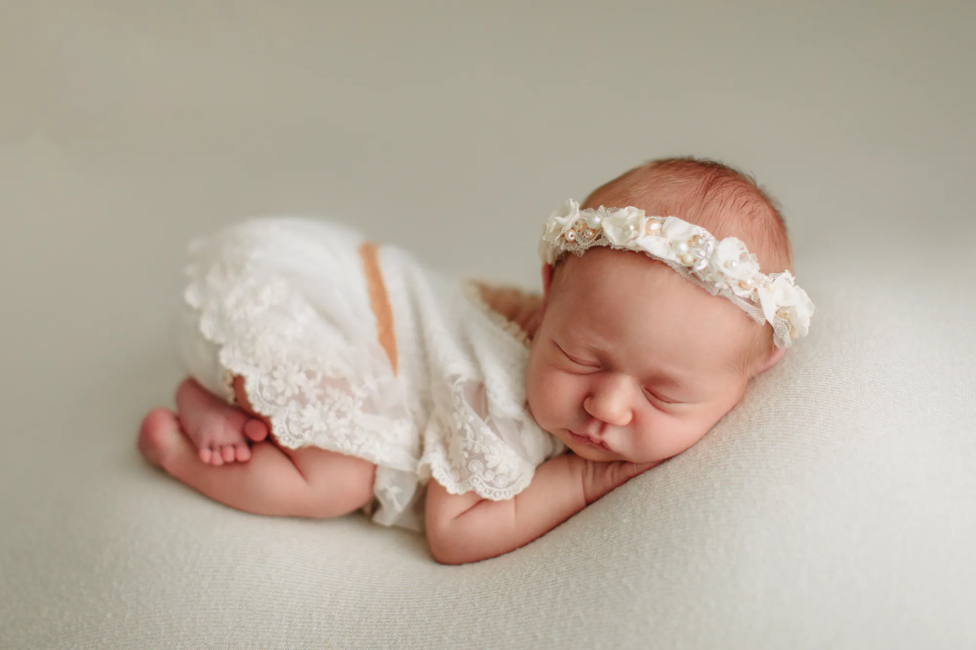 Newborn baby in a lace romper curled up asleep on a cream backdrop