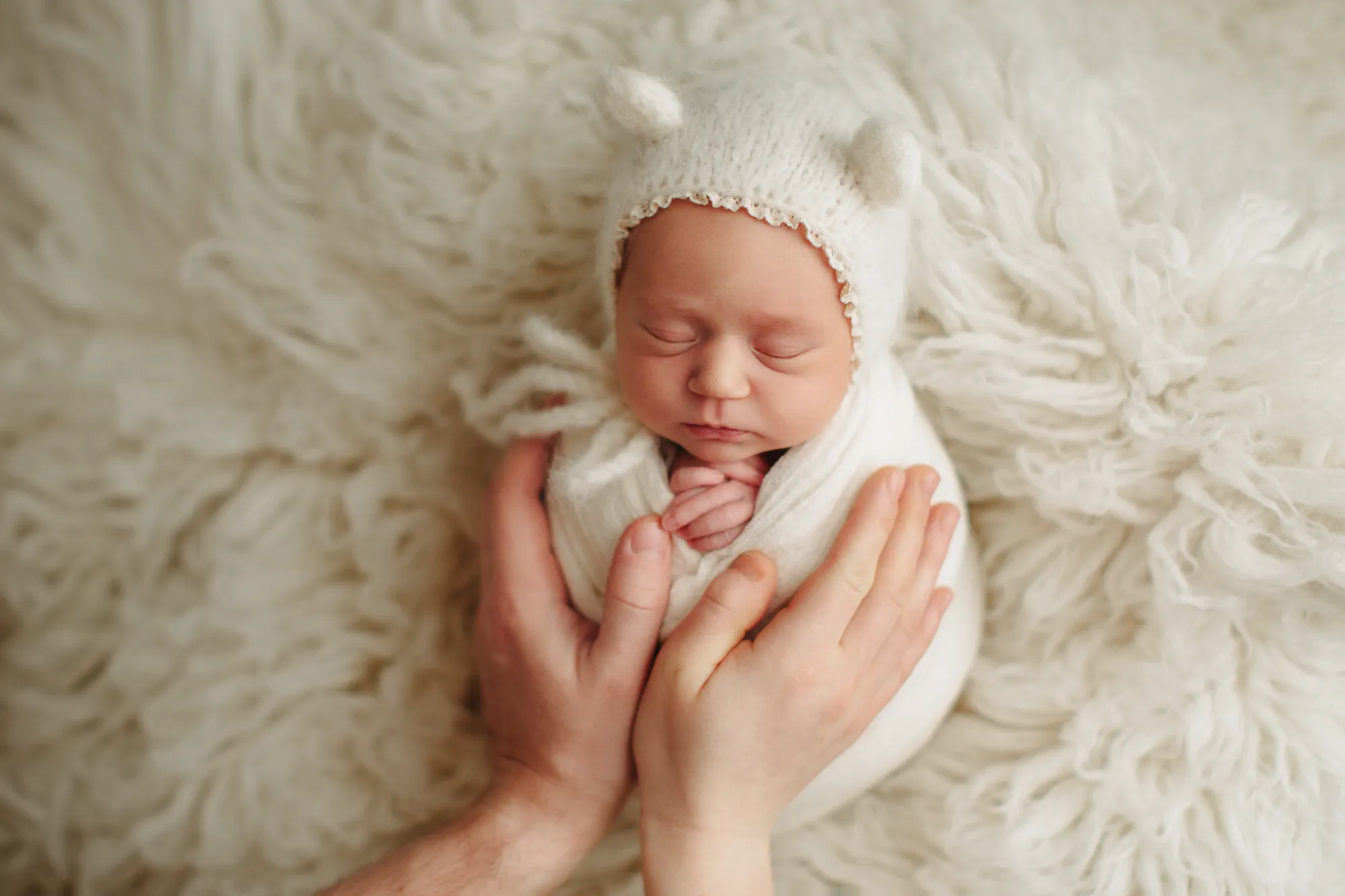 Newborn in a bear-ear bonnet cradled in hands on cream fur