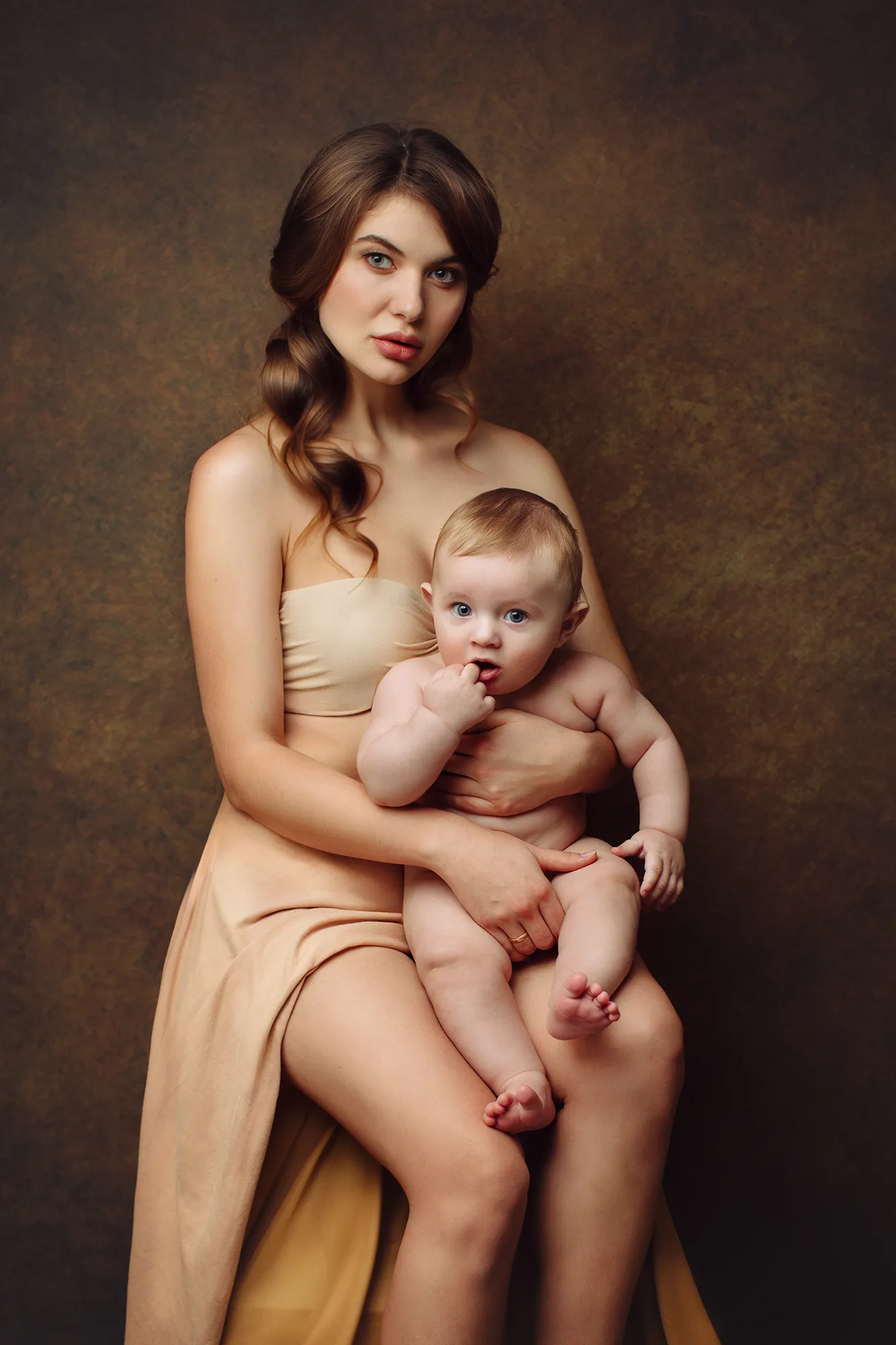 Mother in a nude dress cradling her baby son against a dark backdrop