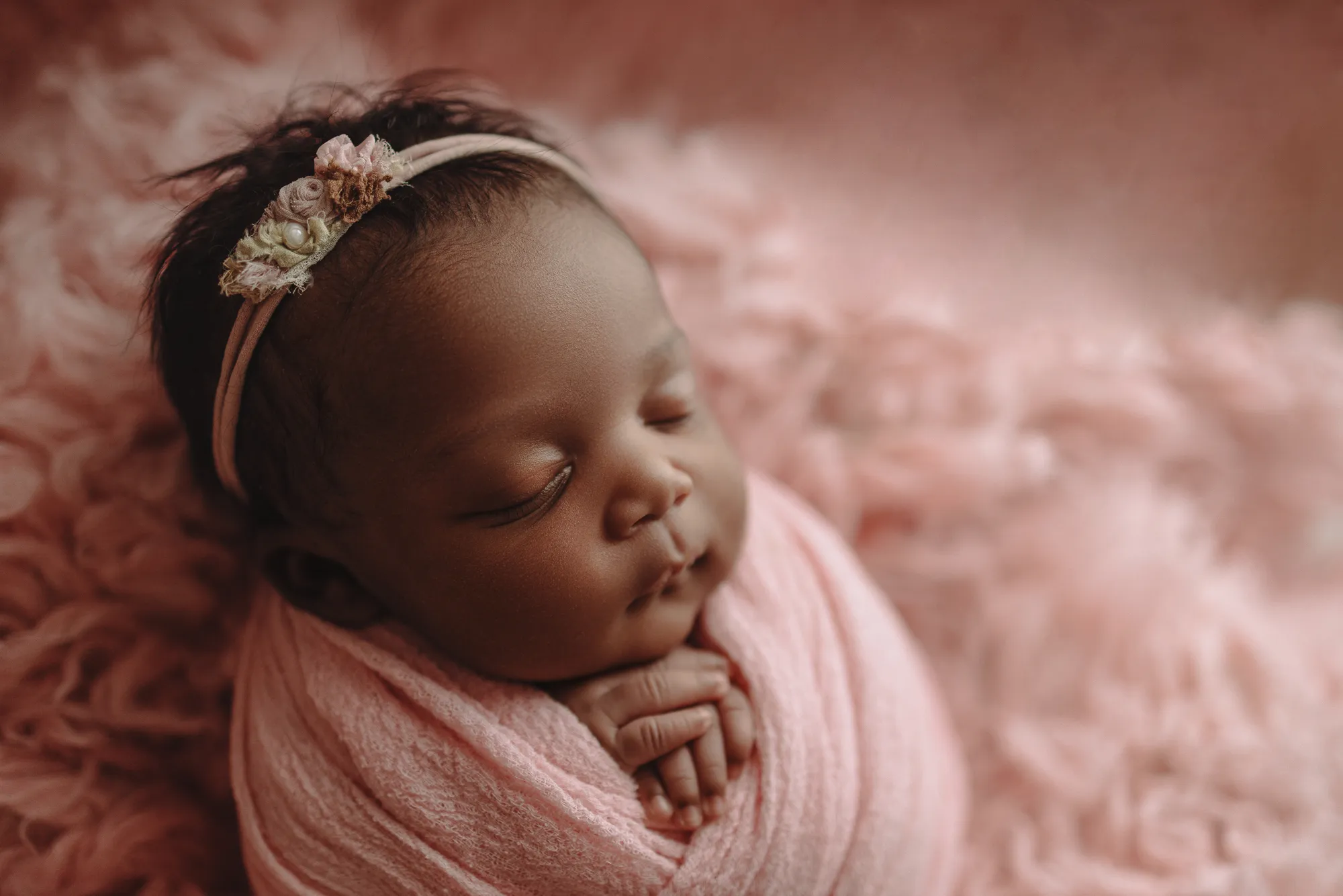 Newborn with a floral headband swaddled in pink on pink fur