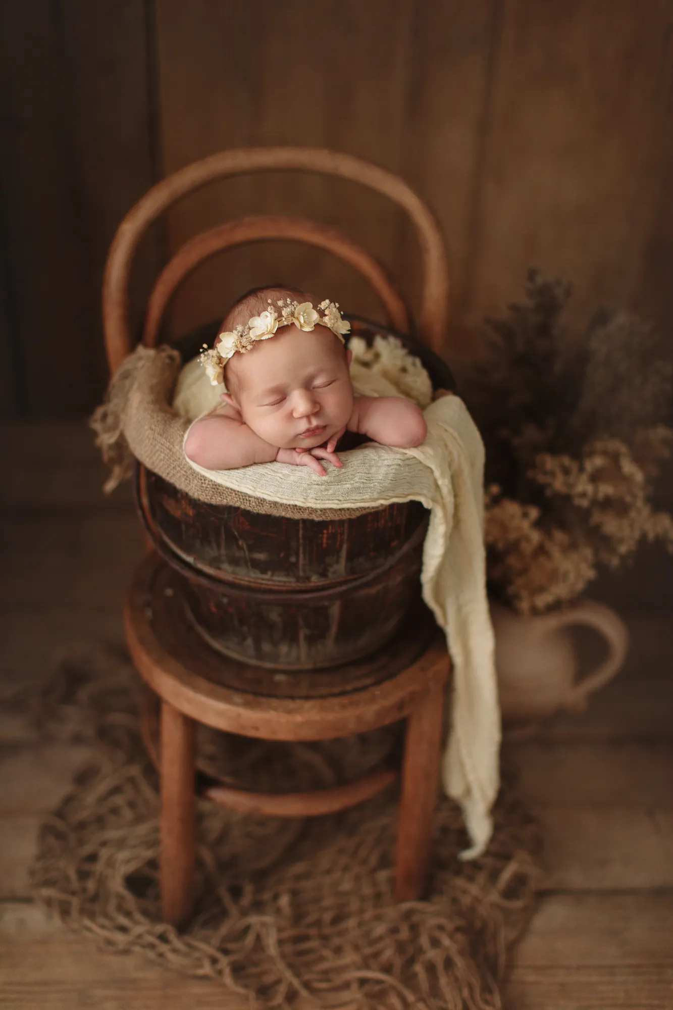 Newborn with a floral headband in a wooden bucket on a vintage chair