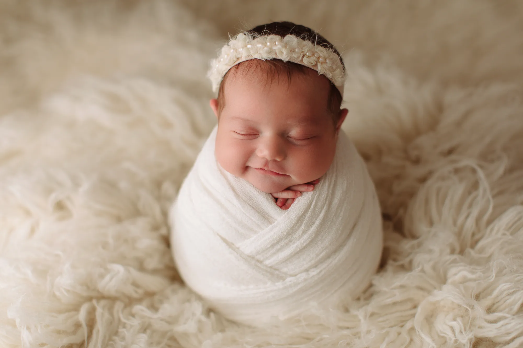 Smiling newborn swaddled in white with a floral headband on cream fur