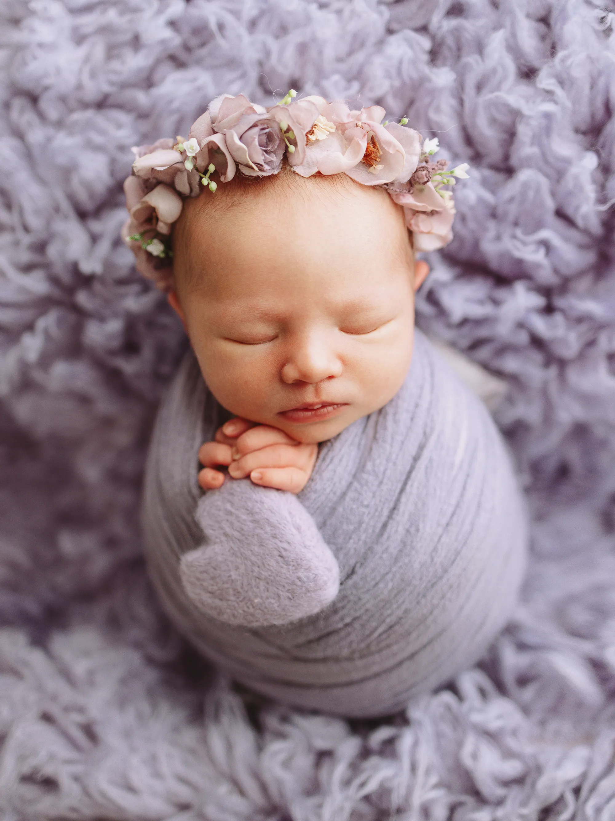 Newborn swaddled in lilac with a felt heart and a flower crown, on lilac fur