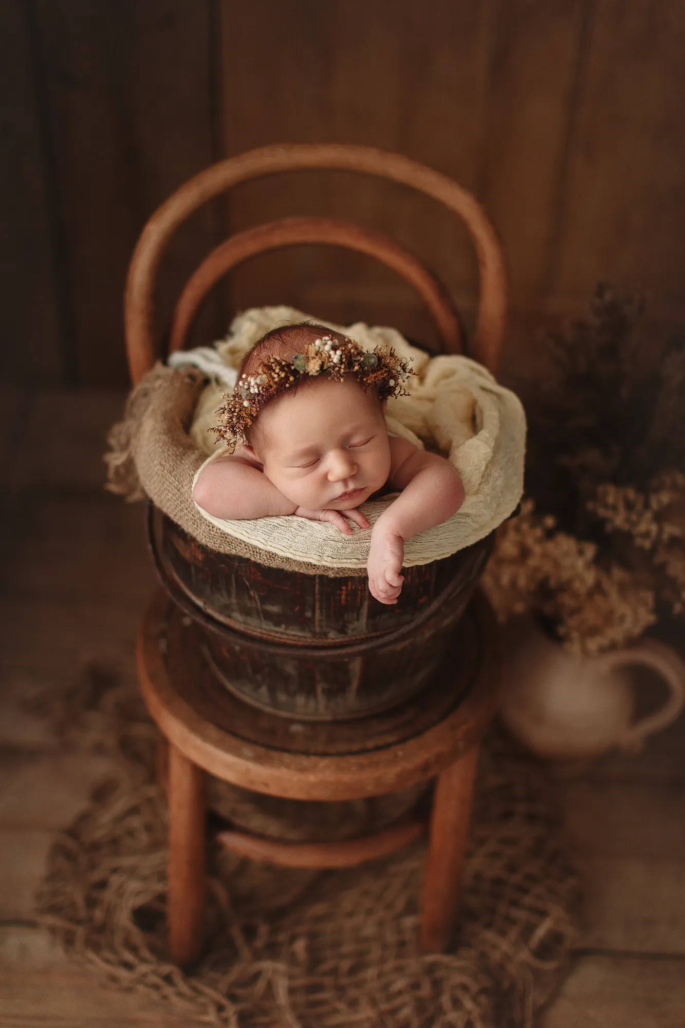 Newborn with a dried-flower crown resting on arms in a bucket on a chair