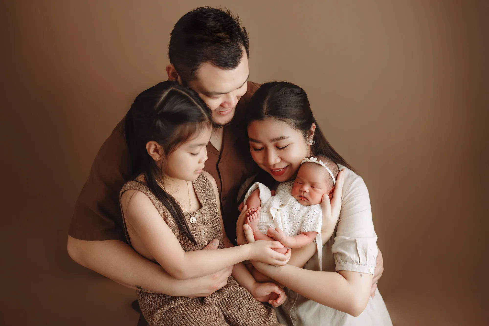 Family with a young daughter holding their newborn baby against a brown backdrop