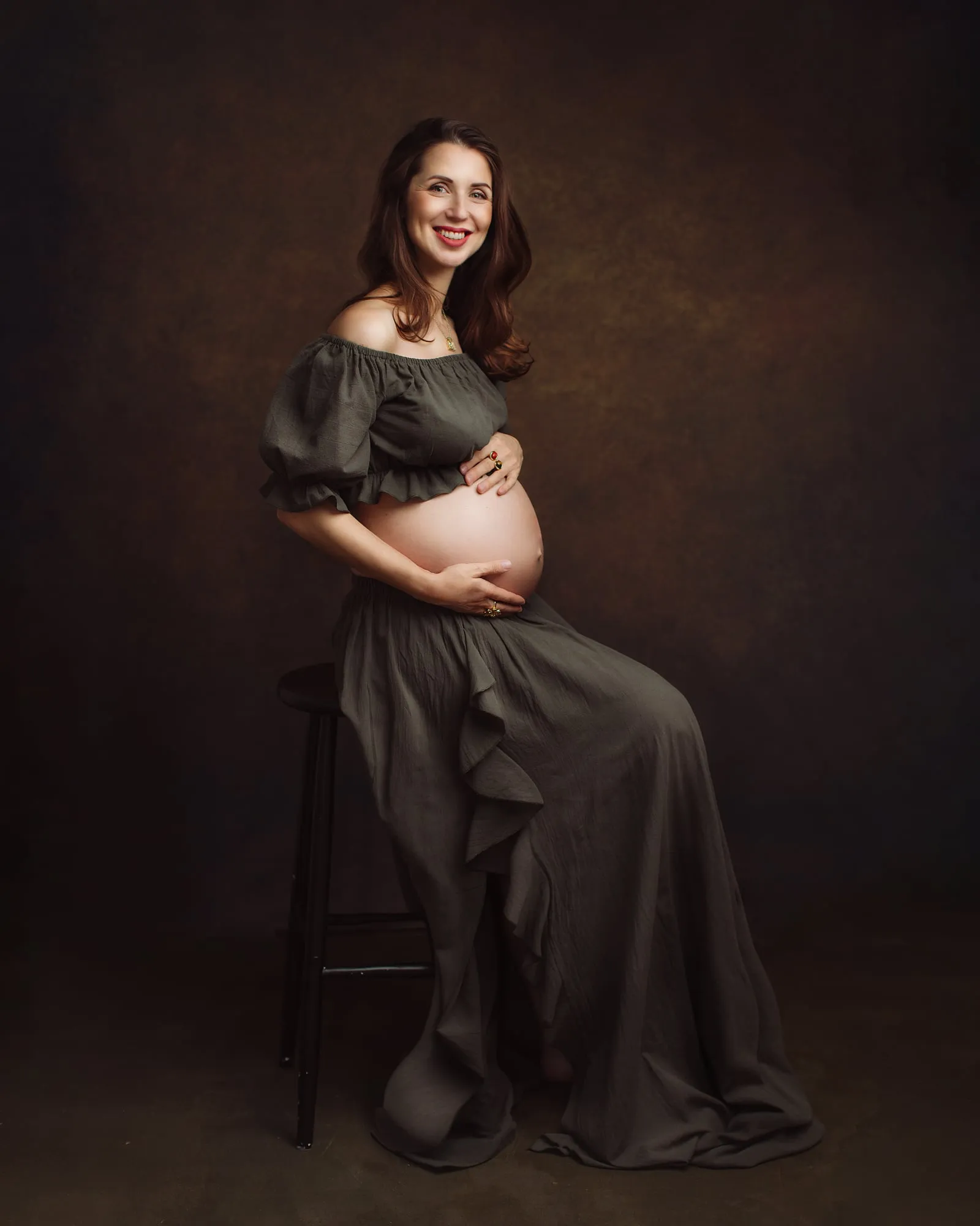 Pregnant woman in an olive off-shoulder dress sitting on a stool in the studio