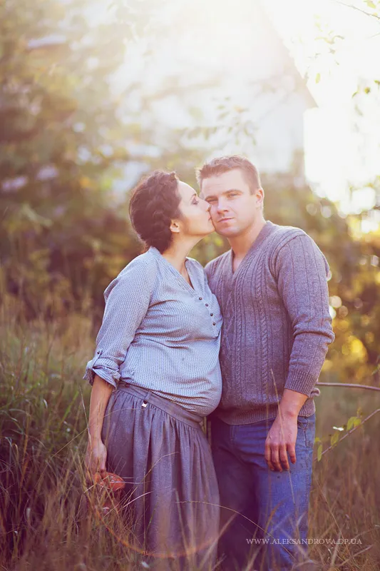 Expectant couple kissing in tall grass at sunset