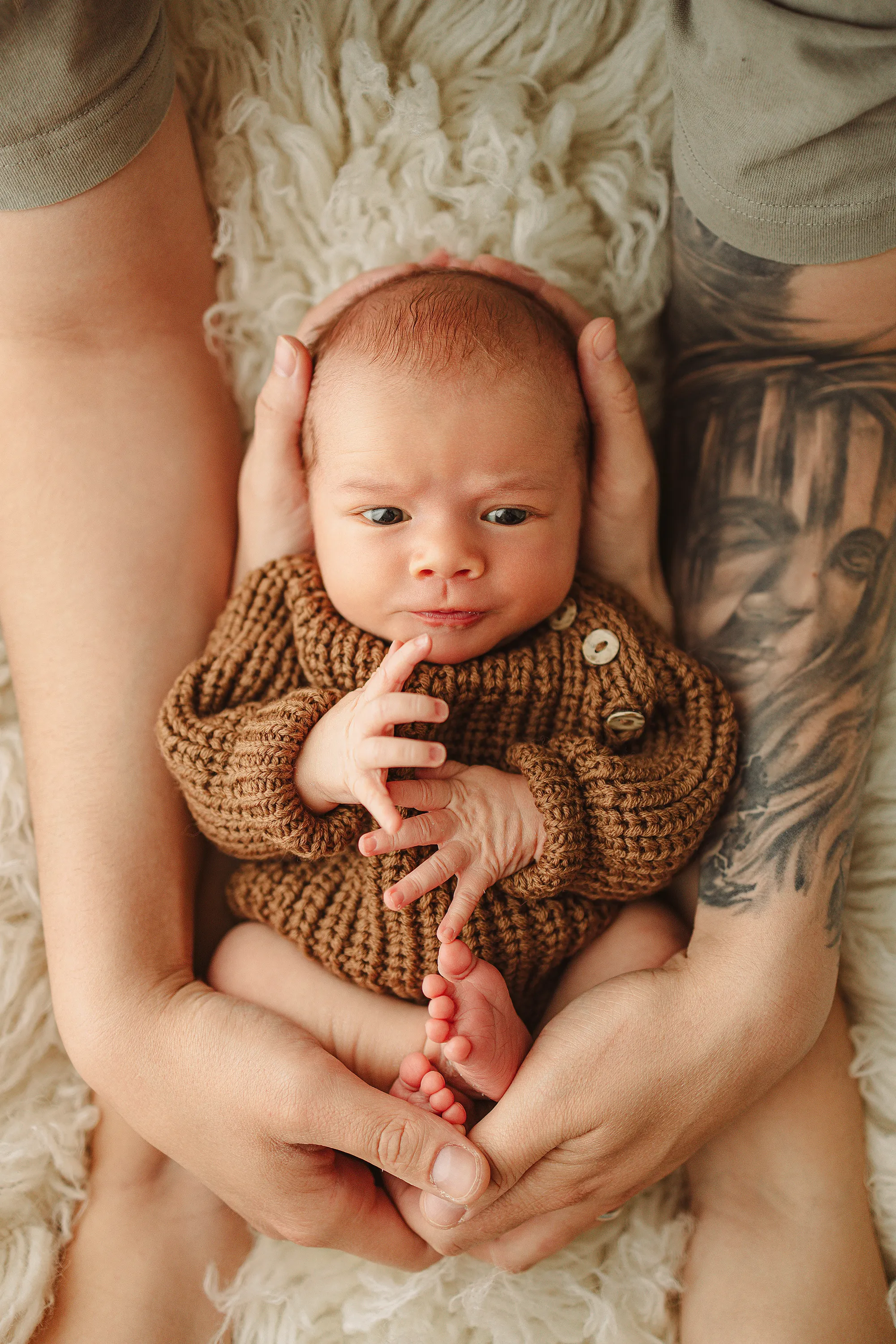 Newborn in a brown knit jumper held in parent's tattooed hands on cream fur