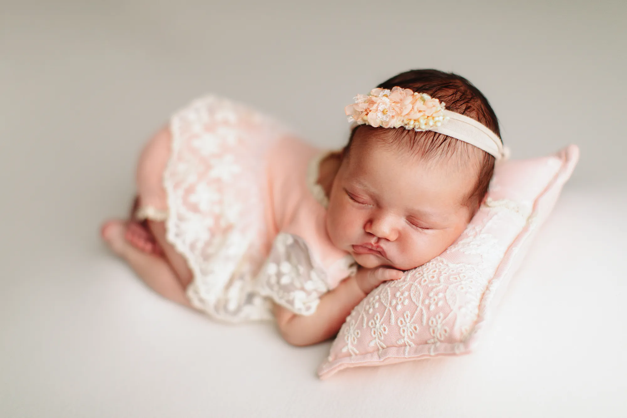 Newborn in a peach lace romper and floral headband asleep on a pillow
