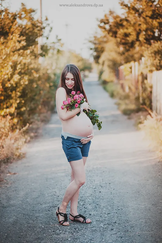 Pregnant woman in denim shorts holding pink roses on a country lane