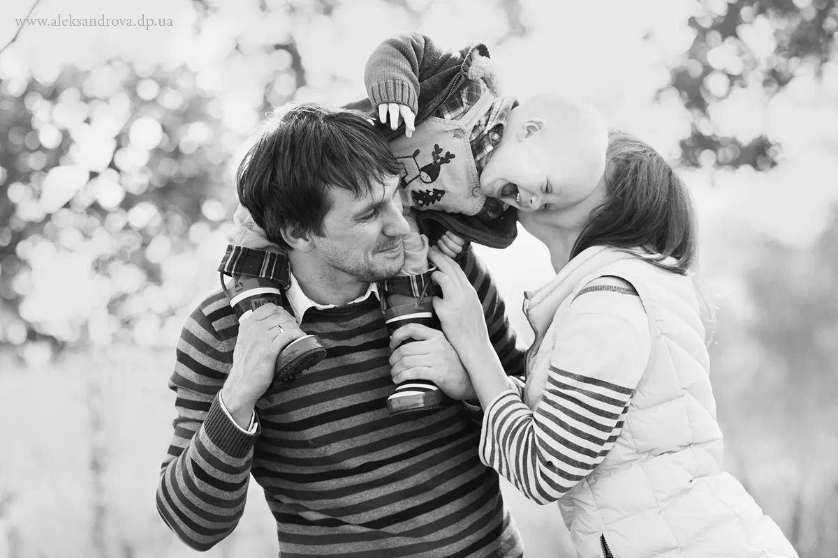 Black and white photo of a dad carrying his son on his shoulders, mum kissing the boy