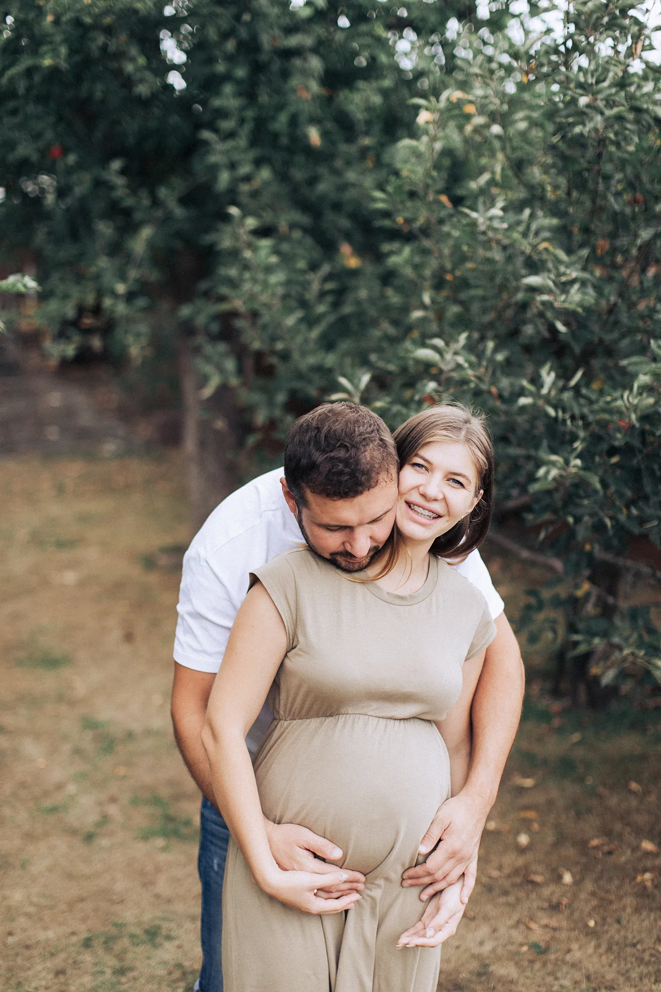Husband hugging his pregnant wife from behind in an orchard