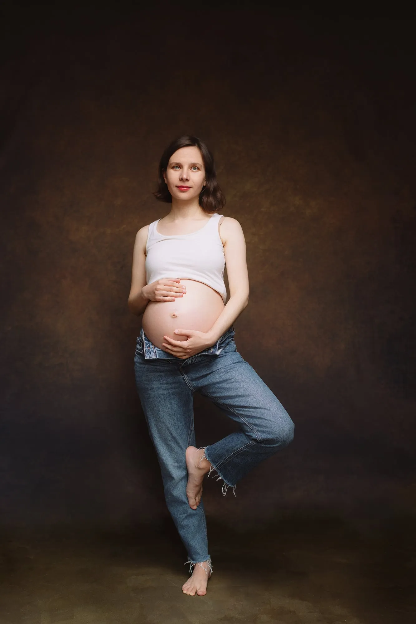 Pregnant woman in a white crop top and jeans, hands on her bump, studio portrait