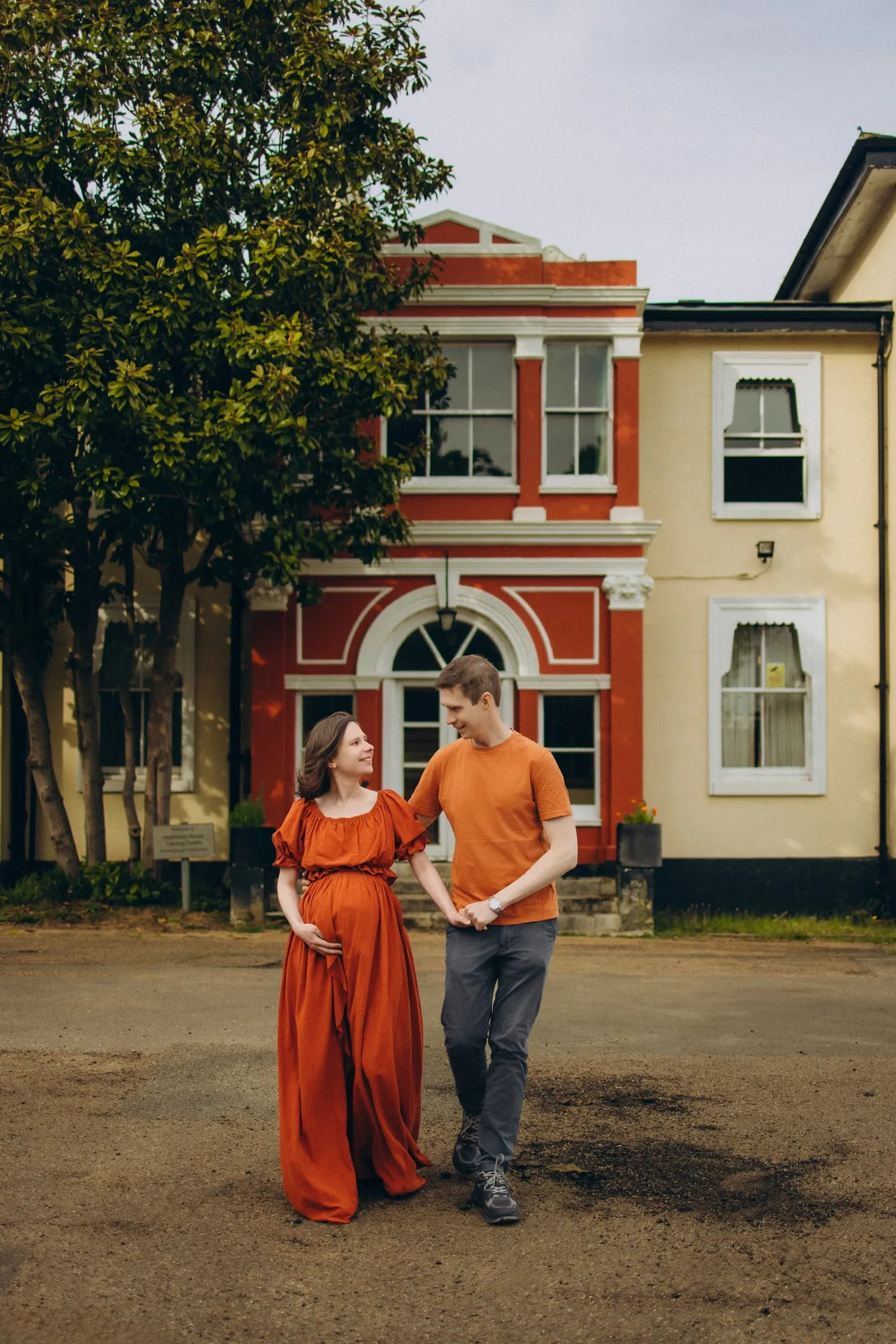 Pregnant woman in an orange dress walking with her partner past a red house