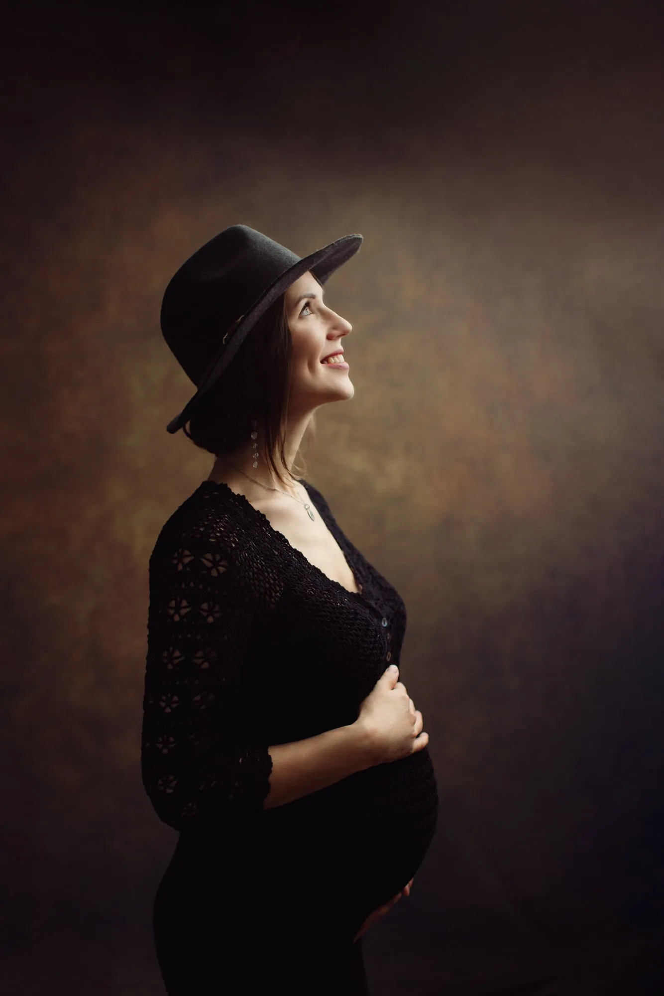 Pregnant woman in a black hat and lace dress smiling upwards, studio portrait