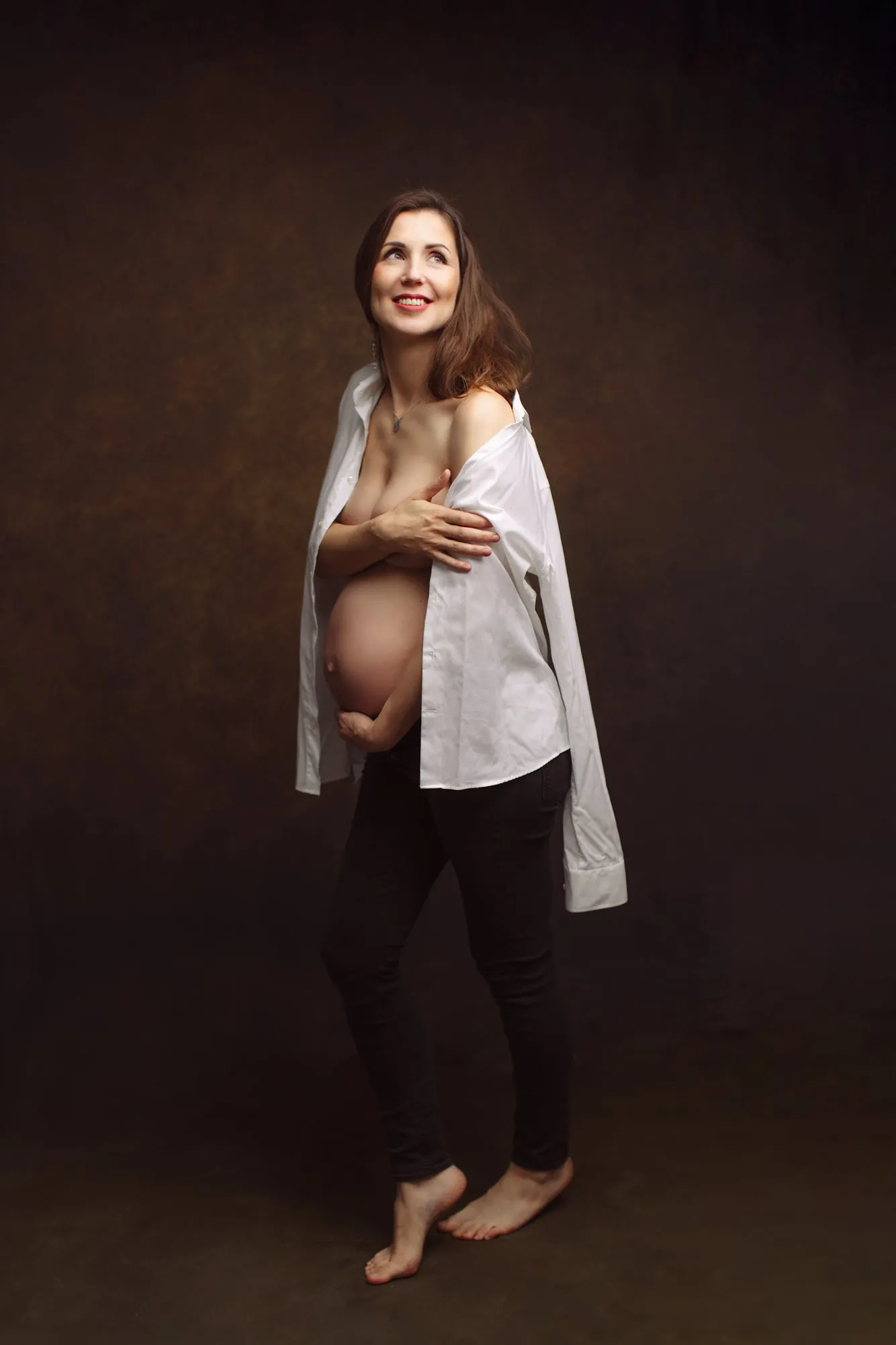 Pregnant woman in a white shirt and black jeans cradling her bump, studio portrait