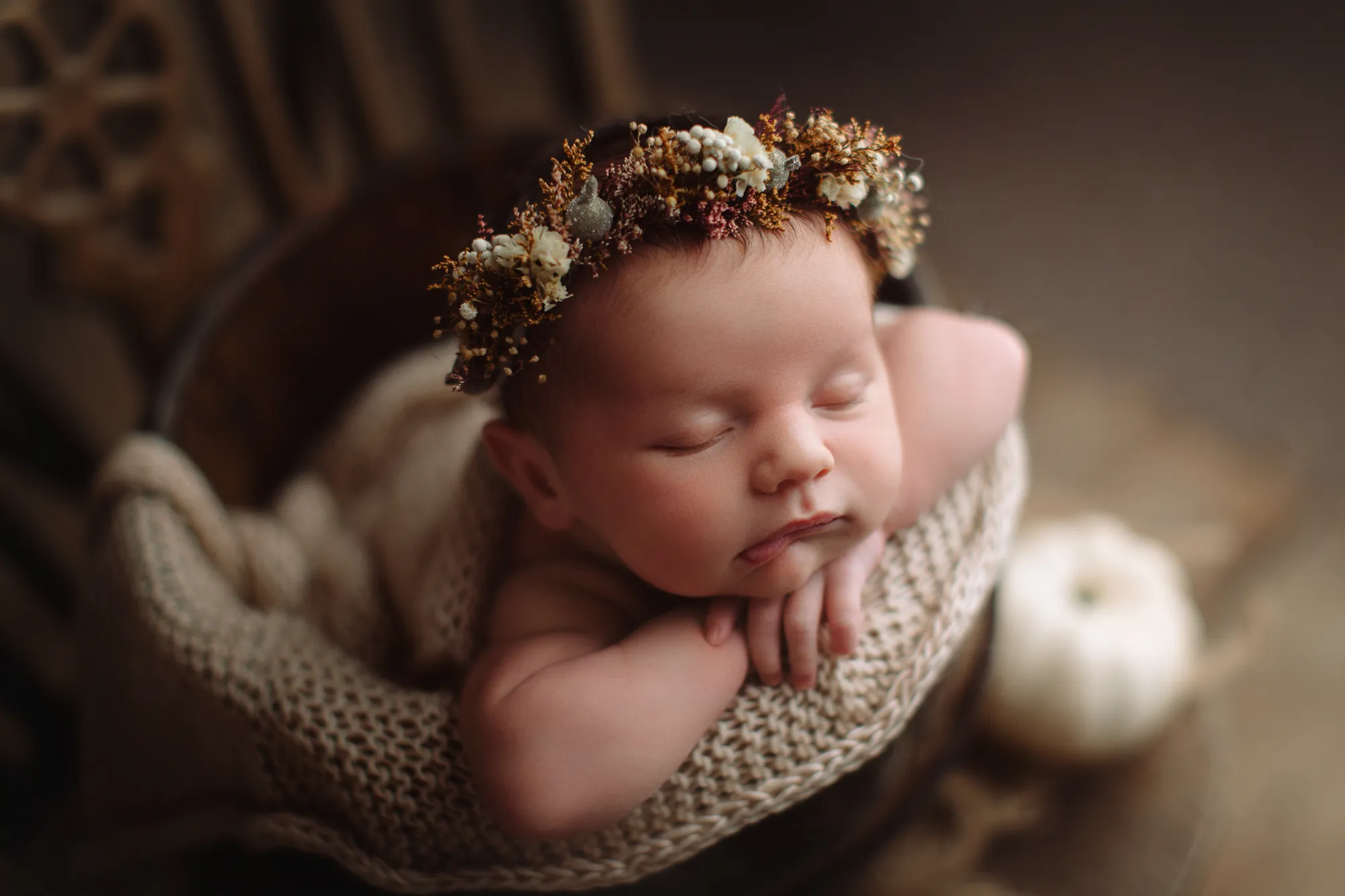 Newborn with a dried-flower crown resting on arms in a knitted bowl