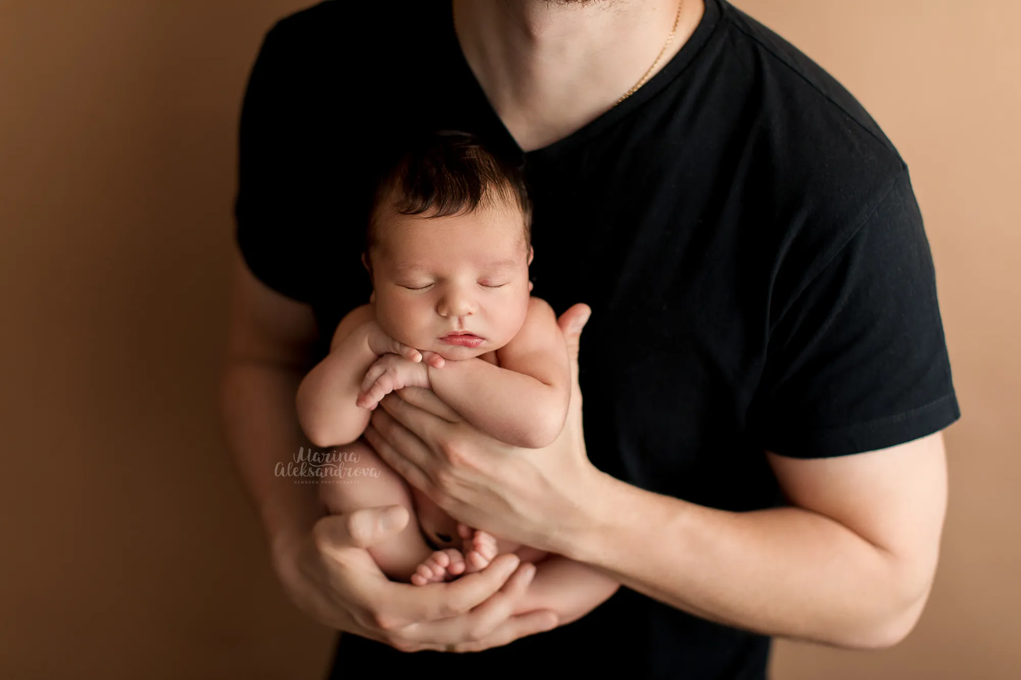 Father in a black T-shirt cradling his sleeping newborn in his hands