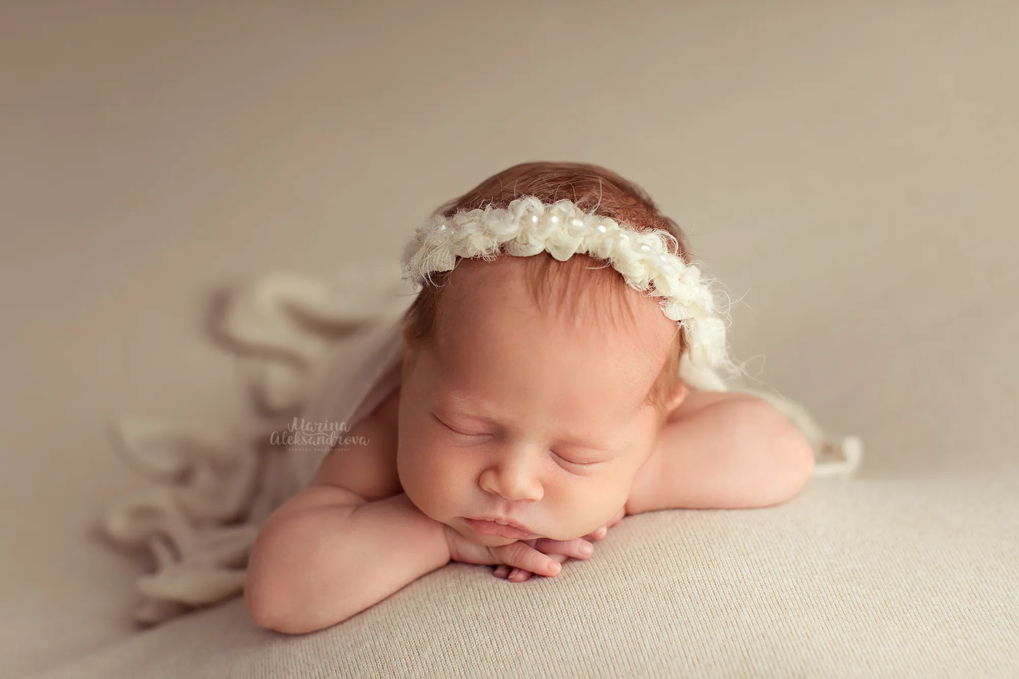 Newborn with a pearl headband resting chin on hands on a beige backdrop