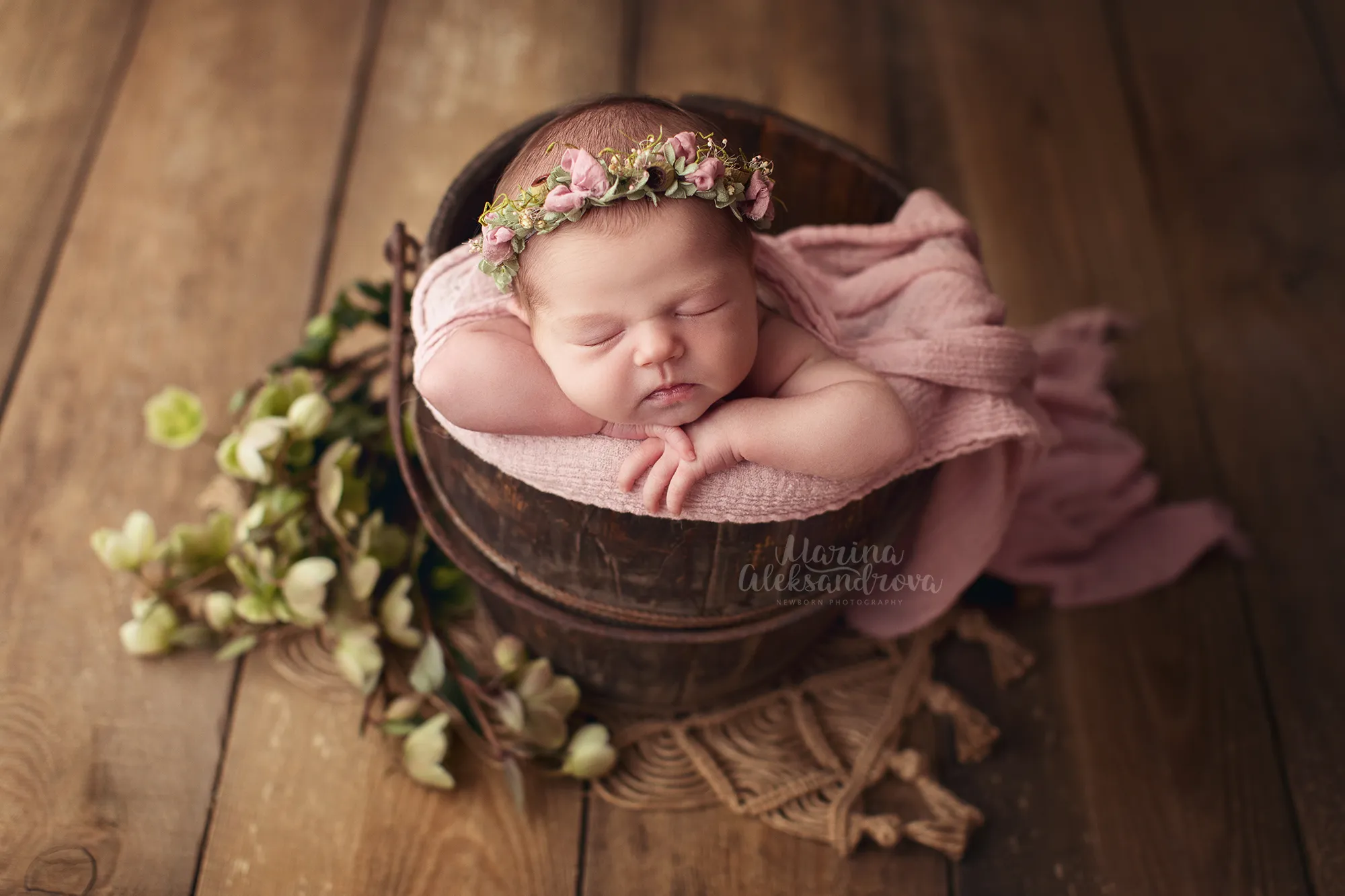 Newborn with a floral headband asleep in a wooden bowl with greenery