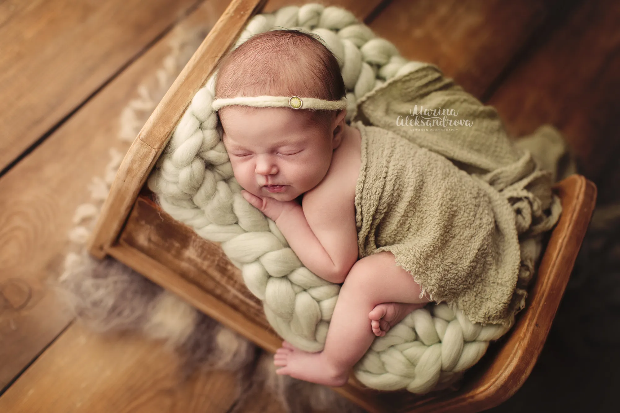 Newborn asleep on a sage chunky-knit blanket in a small wooden bed