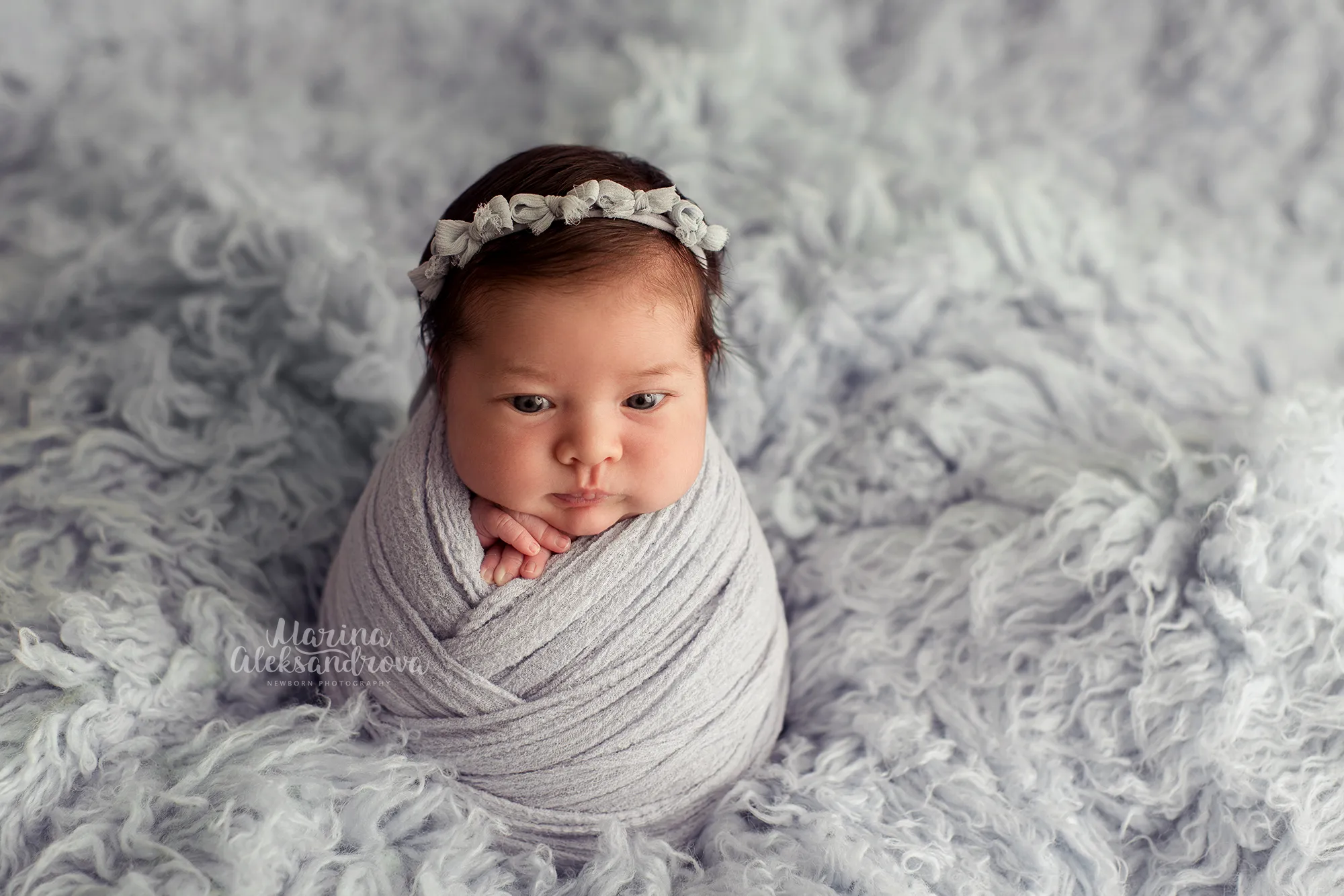 Wide-eyed newborn swaddled in grey with a floral headband on grey fur