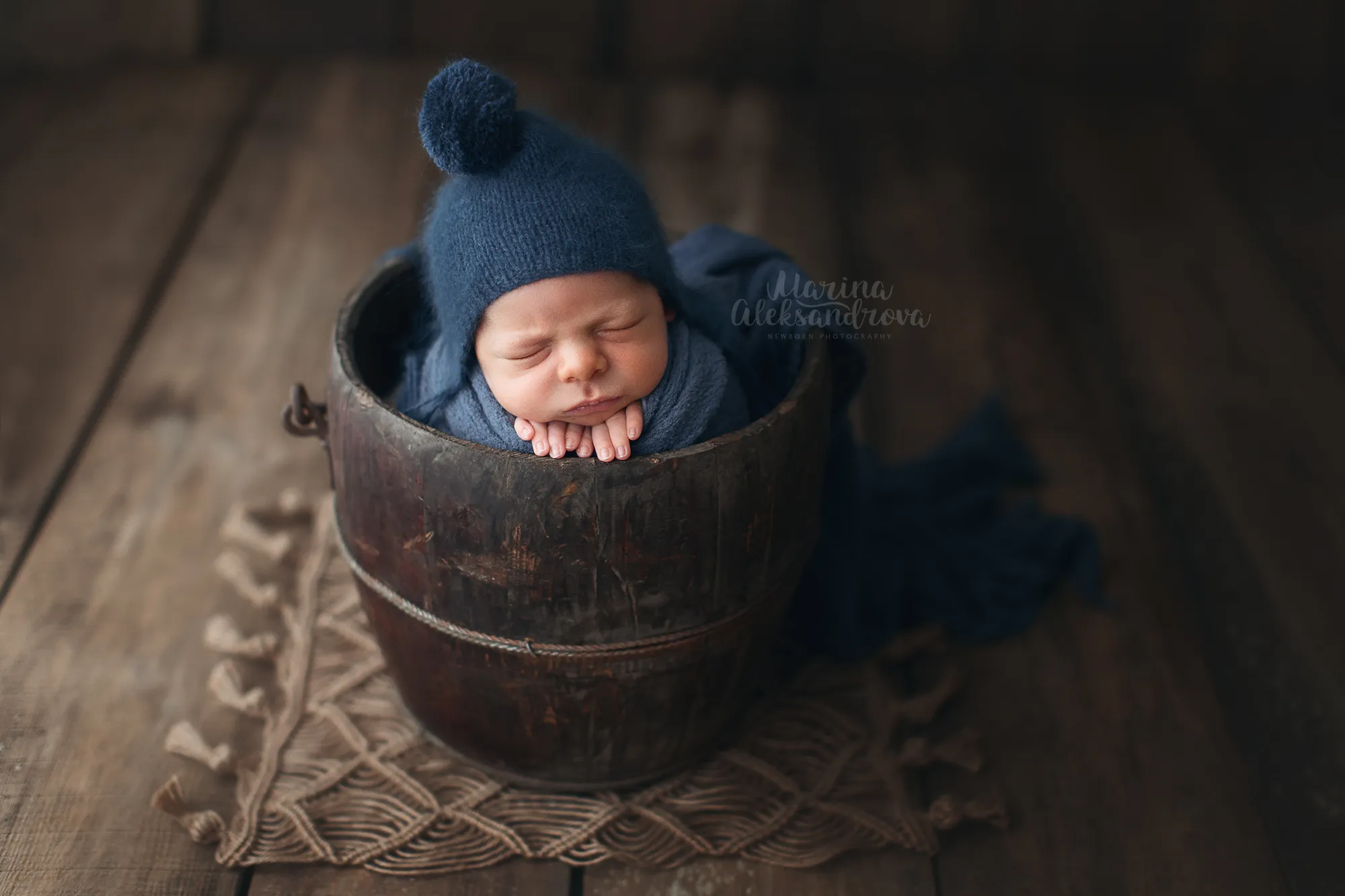 Newborn in a navy pom-pom hat asleep in a wooden bucket