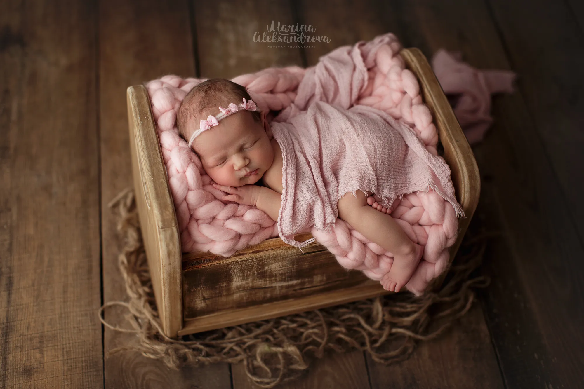 Newborn with a floral headband asleep on a pink knit blanket in a wooden crate