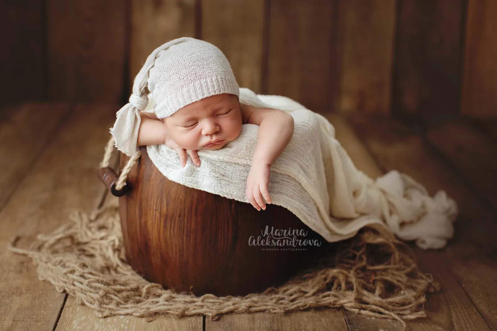 Newborn in a white sleepy hat draped over a wooden bucket